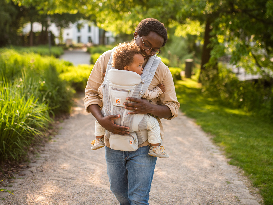 Vater trägt sein Kind in einer Babytrage bei Sonnenschein durch einen Park.