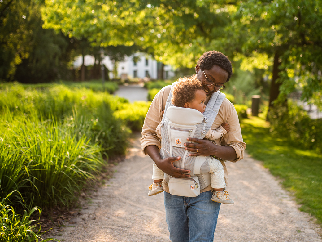 Vater trägt sein Kind in einer Babytrage bei Sonnenschein durch einen Park.