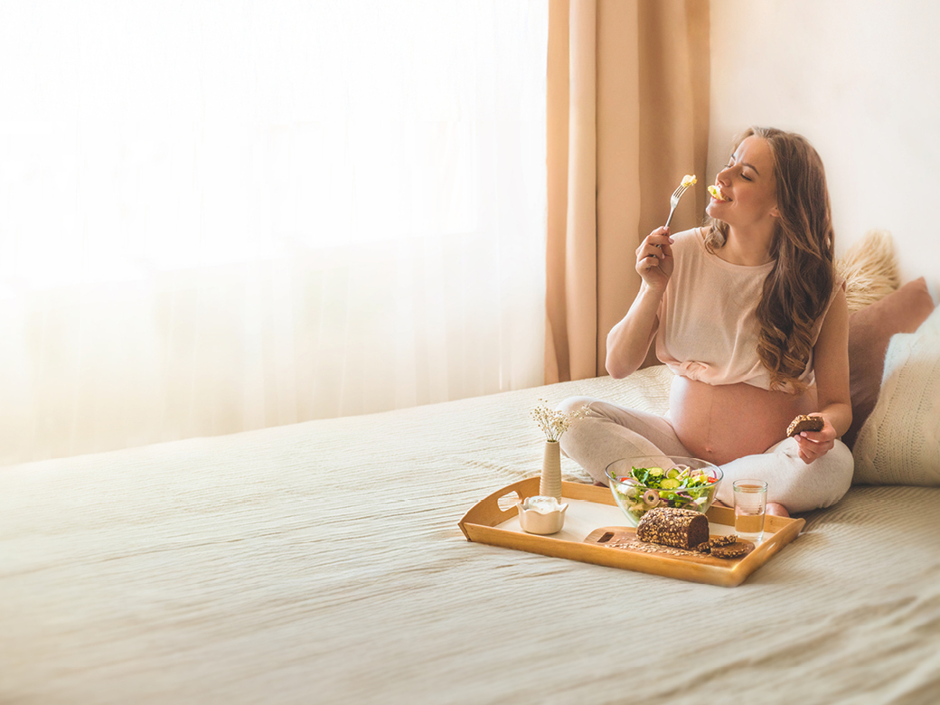 Schwangere Frau sitzt auf dem Bett, hält eine Gabel in der Hand und isst. Vor ihr steht ein Tablett mit Essen.